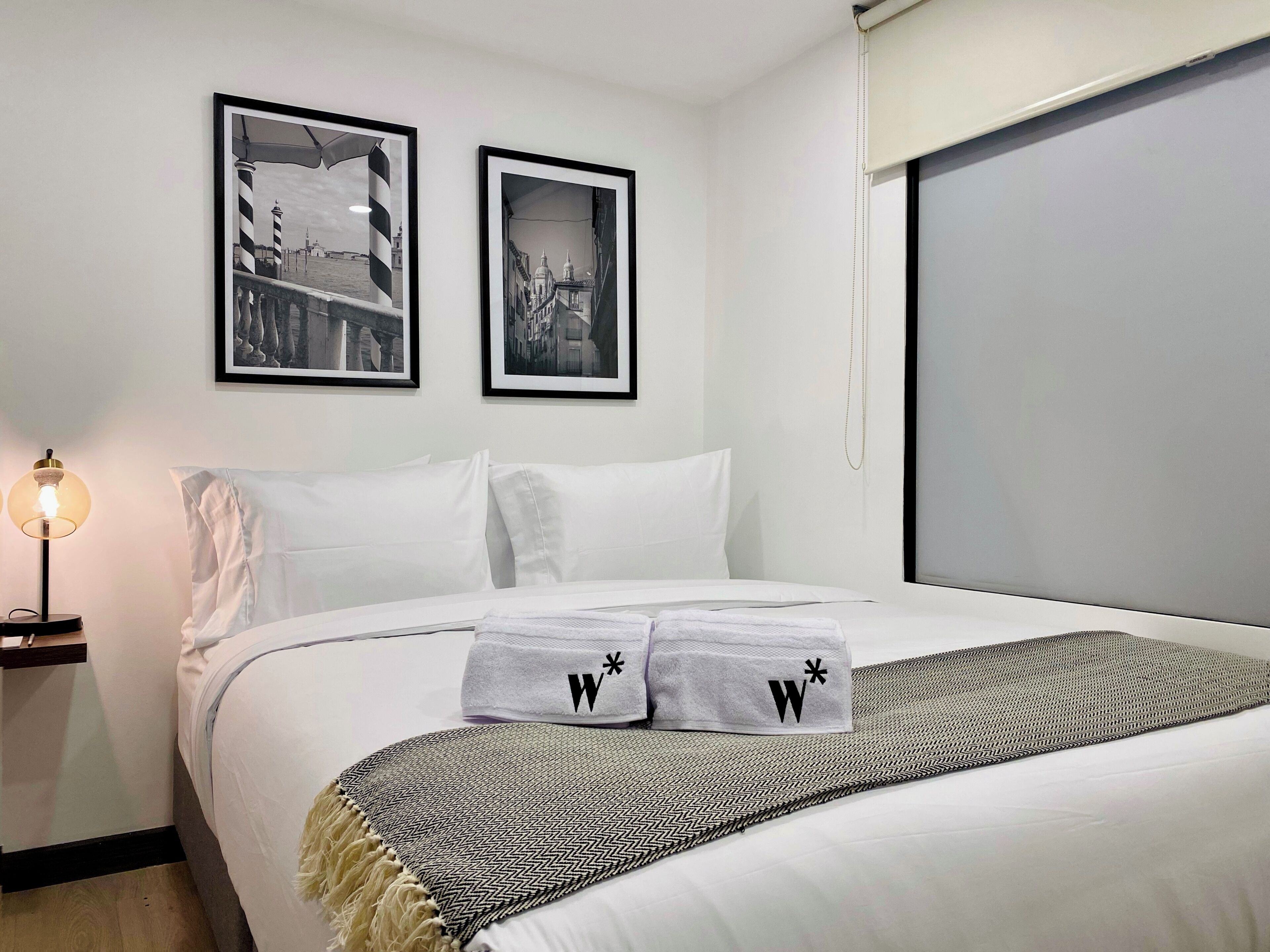 A hotel room with a bed, two framed black and white photographs on the wall, a bedside lamp, and a window with a blind.