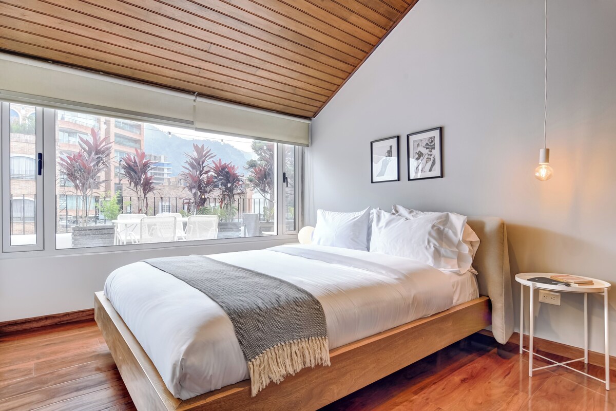 Ein modernes Schlafzimmer mit Holzbalkendecke, großen Fenstern, einem Bett mit weißer Bettwäsche und einem kleinen Nachttisch.