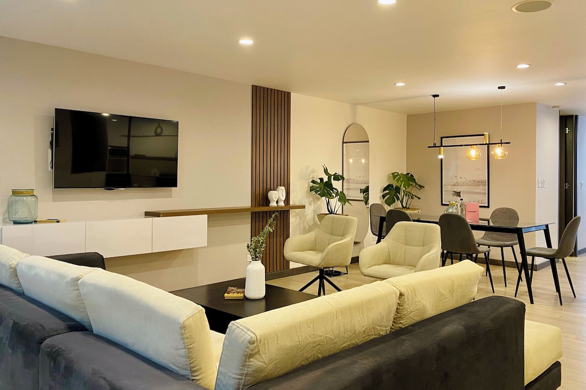Ein modernes Wohnzimmer mit Sofa, Sesseln, einem Couchtisch, einem Fernseher und einem Essbereich mit Tisch und Stühlen.