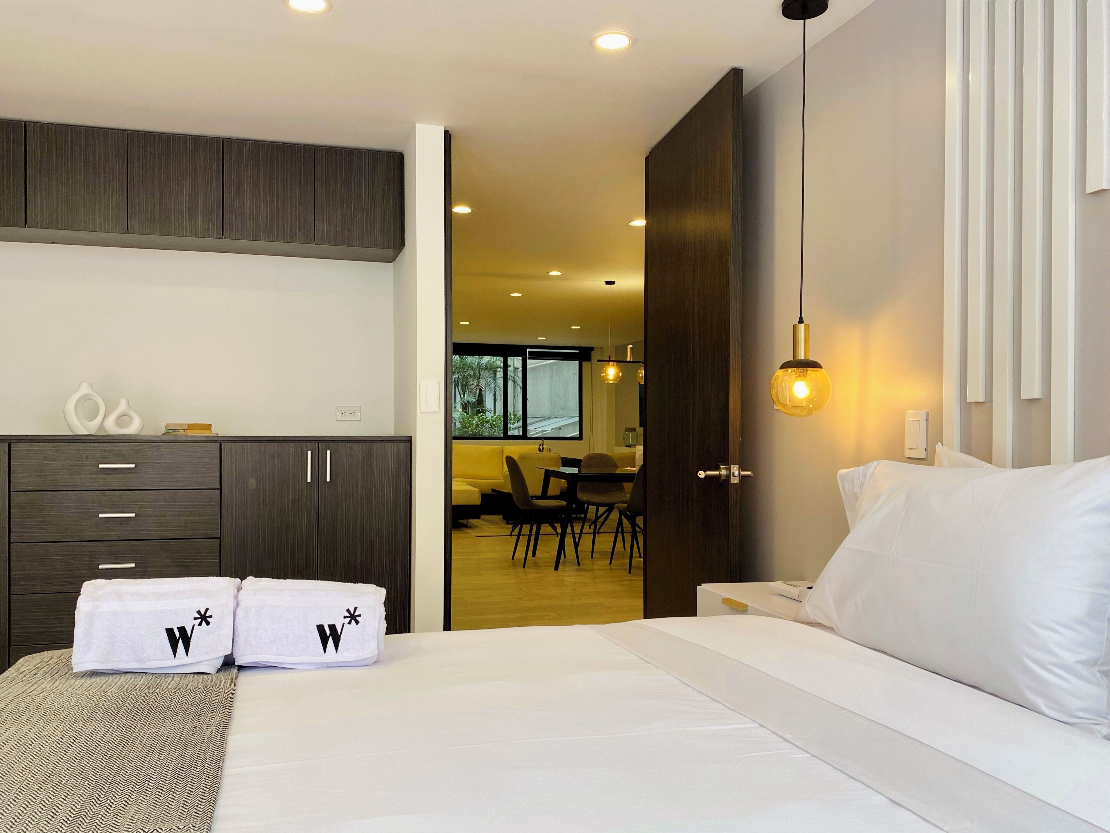 Une chambre d’hôtel moderne, équipée d’un lit, d’une armoire et d’un coin repas, le tout visible à travers une porte ouverte.