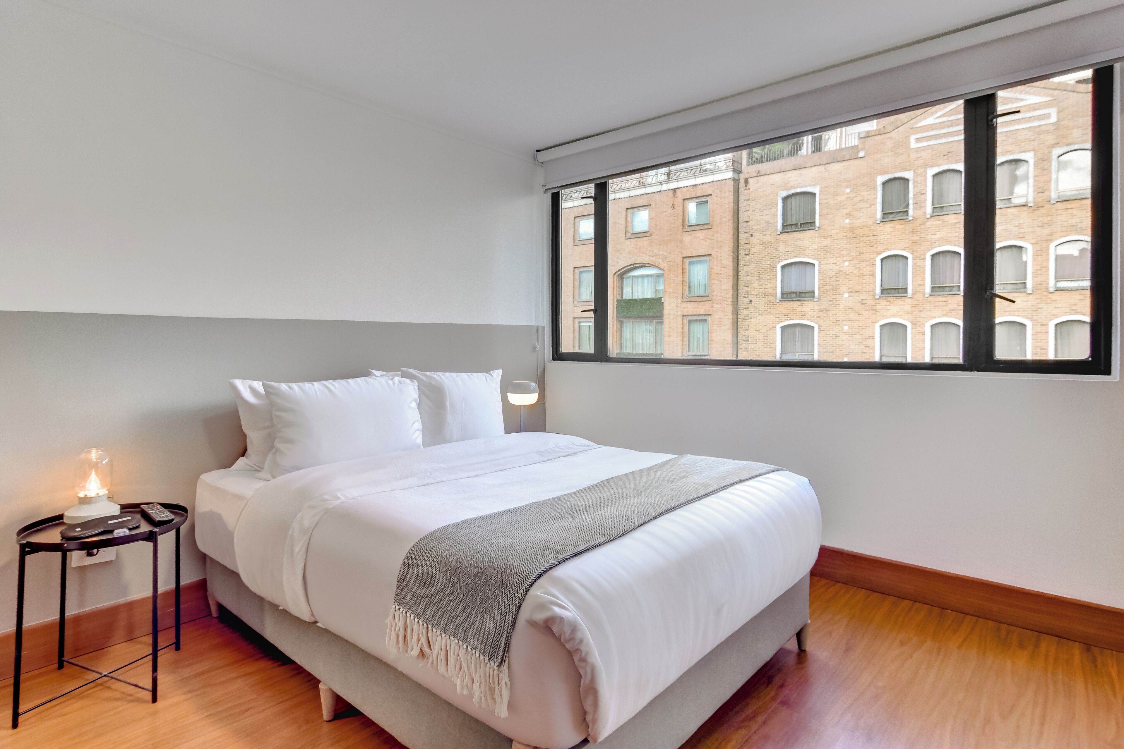 Ein ordentlich gemachtes Bett mit weißer Decke und grauer Tagesdecke, ein Nachttisch mit Lampe sowie ein großes Fenster mit Blick auf ein Backsteingebäude.