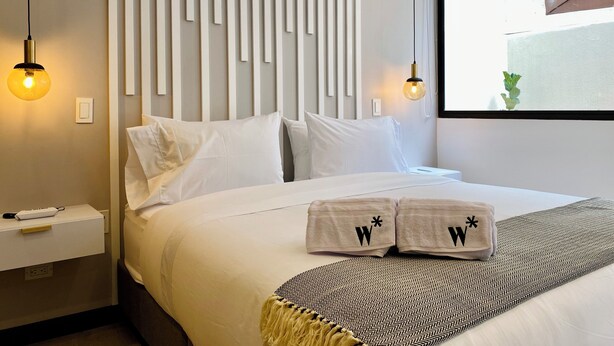 Una habitación de hotel moderna con una cama grande, ropa blanca, una manta con estampado y una lámpara fijada a la pared.