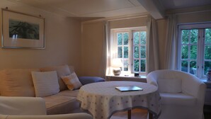 Una habitación con sofá, dos sillones, una mesa redonda con mantel, una lámpara y una ventana con cortinas.