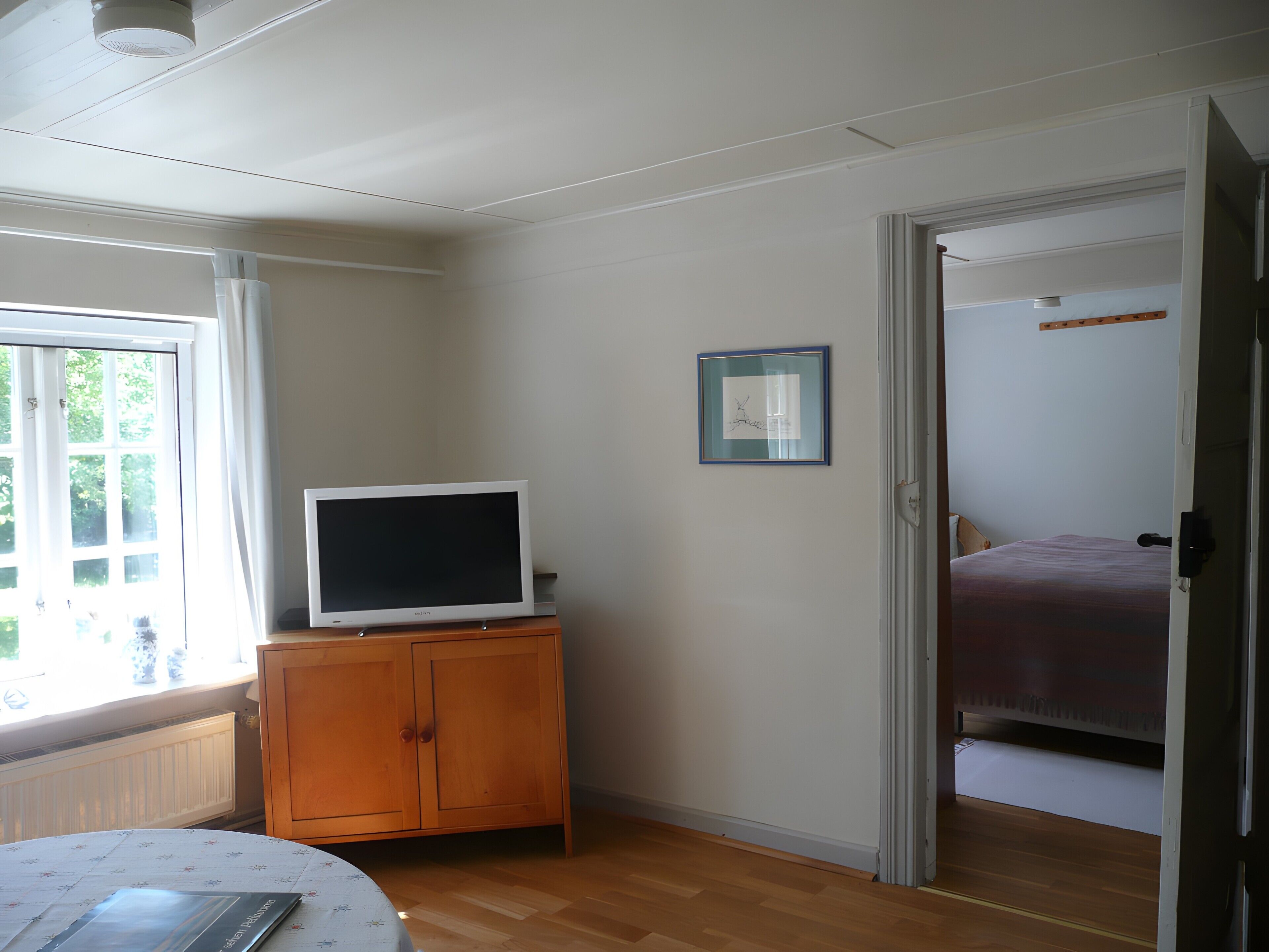 Ein Zimmer mit einem Holzkasten, einem Fernseher, einem gerahmten Bild an der Wand und einem Bett im angrenzenden Raum.