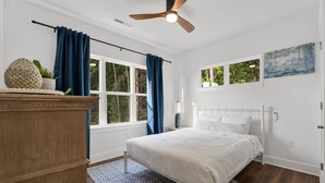 Un dormitorio con cama, mesita de noche, ventilador de techo, ventana con cortinas, cómoda de madera y un cuadro en la pared.