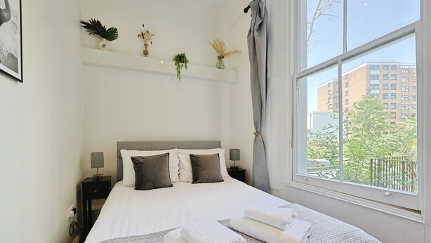 Cama bem arrumada com lençóis brancos e colcha bege, mesa de cabeceira com abajur, janela com vista para edifícios e vegetação, e prateleira com objetos decorativos.