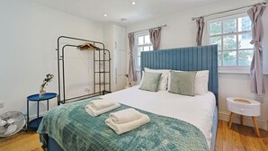 Um quarto com cama, mesa de cabeceira, ventoinha, janela com cortinas e guarda-roupa.