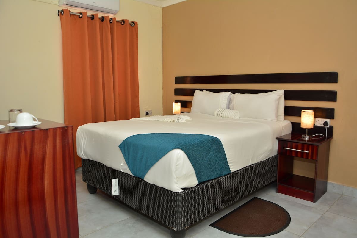 A hotel room with a bed, a nightstand with a lamp, a wooden desk with a cup, and a brown rug.