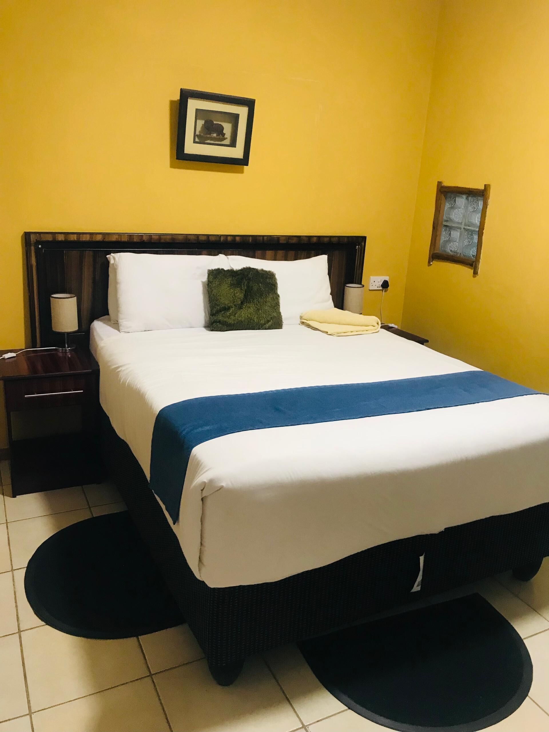 A hotel room with a bed, a nightstand, a lamp, a small window, and a framed picture on the wall.