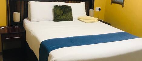 A hotel room with a bed, a nightstand, a lamp, a small window, and a framed picture on the wall.