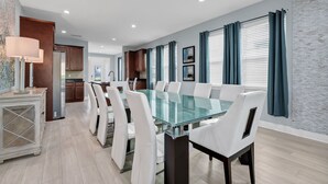 Un comedor moderno con una mesa de vidrio y sillas blancas, una cocina con armarios de madera y cortinas color turquesa en las ventanas.