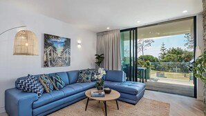 A modern living room with a blue sectional sofa, a coffee table, and a large window offering a view of the outdoors. - Byron Bay Accom - Forty Five on Lawson Apt 7 (Byron Bay)