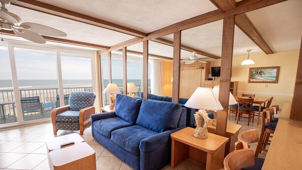 A spacious living area with a blue sofa, wooden furniture, and large windows offering a view of the beach. - Oceanfront Studio w/ Balcony in Daytona Beach (Daytona Beach Shores)