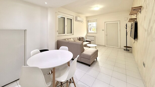 A modern living room with a sofa, a dining table with chairs, a TV mounted on the wall, and a door. - Adriatic Central 2BDR Apartment (Budva)