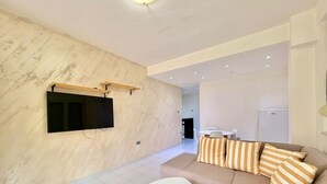 A modern living room with a flat-screen TV mounted on a textured wall, a beige sofa with striped cushions, and a small white table. - Adriatic Central 2BDR Apartment (Budva)
