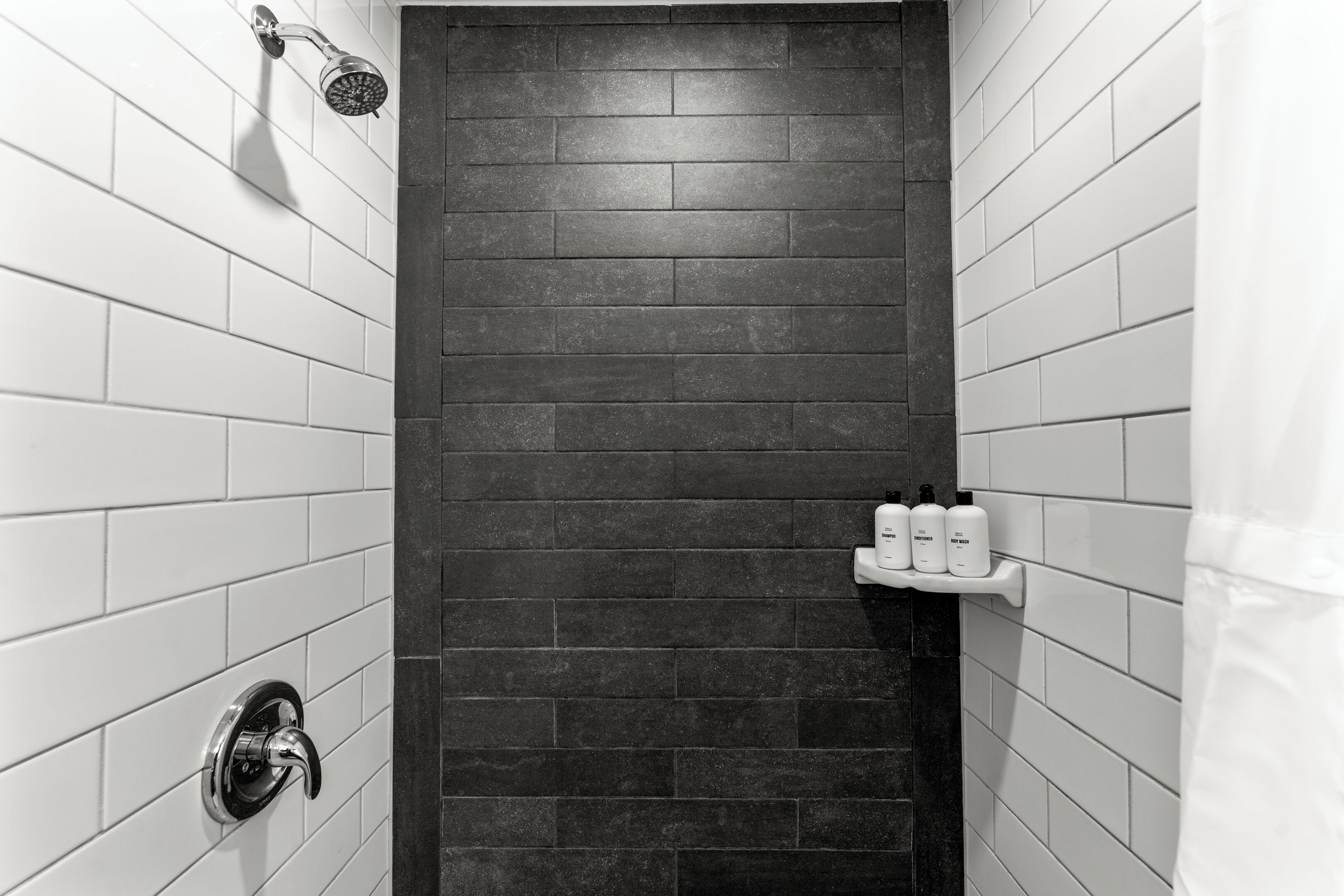 Un baño con azulejos blancos y oscuros, un cabezal de ducha, un grifo y un estante con artículos de aseo.