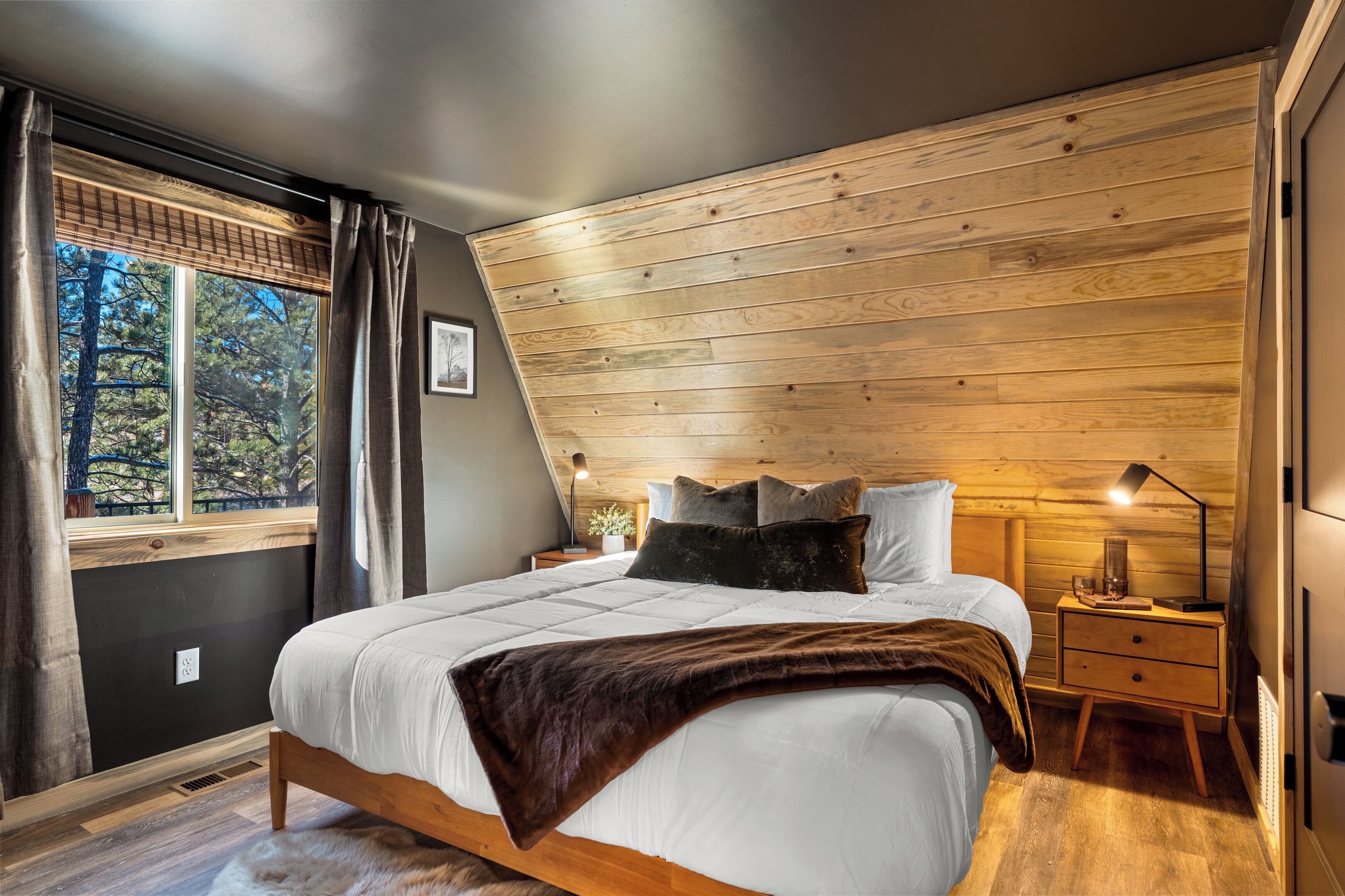 Une chambre avec un plafond en bois, un grand lit recouvert d’une couette blanche, un plaid marron et une table de chevet avec une lampe.