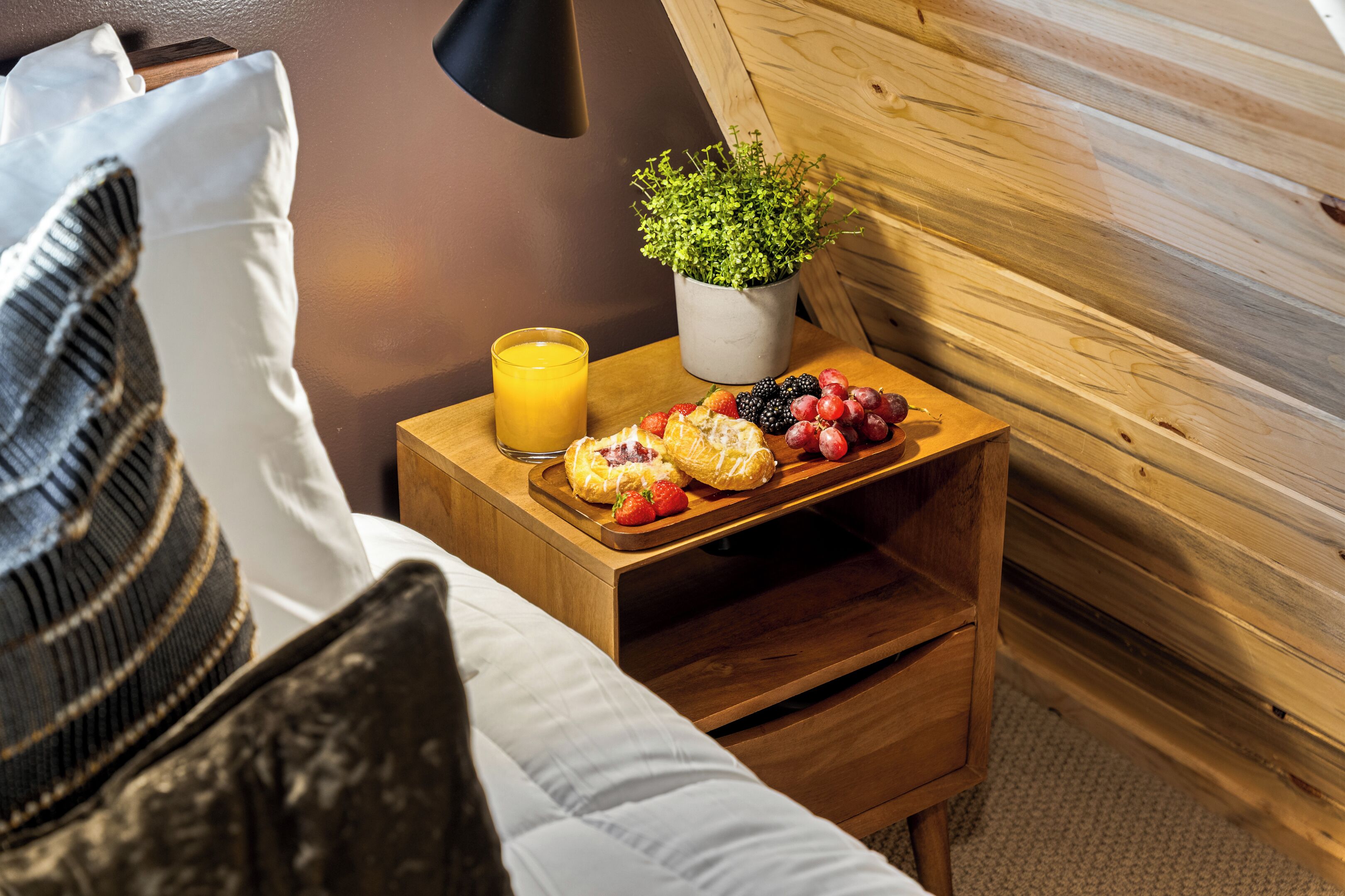 A wooden bedside table with a glass of orange juice, a potted plant, and a plate of fruit and pastries.