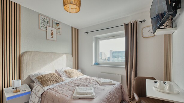 Une chambre d’hôtel moderne équipée d’un lit, d’une table de chevet et d’un bureau avec une chaise. On y trouve une fenêtre avec des rideaux, une horloge et des œuvres d’art accrochées au mur.