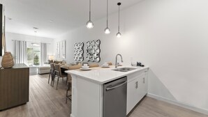 A modern kitchen with a central island, stainless steel appliances, pendant lighting, and a dining area with a view. - Modern 4BR Home + Pool + Parking Disney 15min (Davenport)