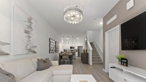 A modern living room with a sectional sofa, a white coffee table, and a chandelier. There is a staircase leading to an upper level. - Modern 4BR Home + Pool + Parking Disney 15min (Davenport)