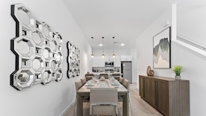 A modern dining area with a long table set for six, a kitchen in the background, and a large abstract wall art piece. - Modern 4BR Home + Pool + Parking Disney 15min (Davenport)