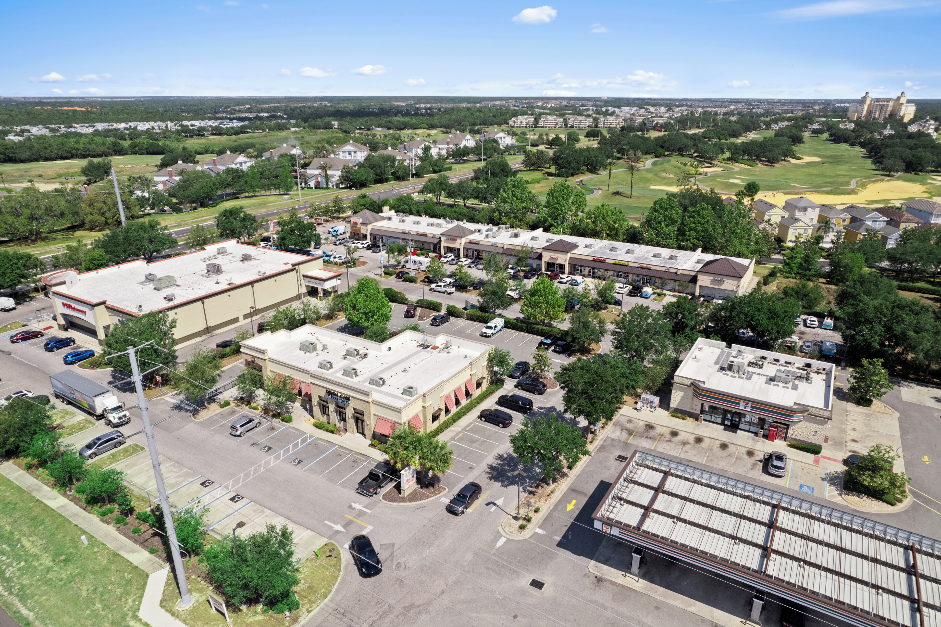 An aerial view of a commercial area with a parking lot, a large building with a flat roof, and surrounding greenery.