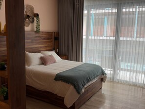 A bedroom with a wooden headboard, a bed with white bedding and a green throw, a nightstand, a wall-mounted plant, and a window with sheer curtains. - Centro Playa Del Carmen Duna  206 (Playa del Carmen)
