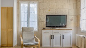 Een kamer met een houten garderobe, een witte kast met lades, een televisie erbovenop, een fauteuil en een radiator.