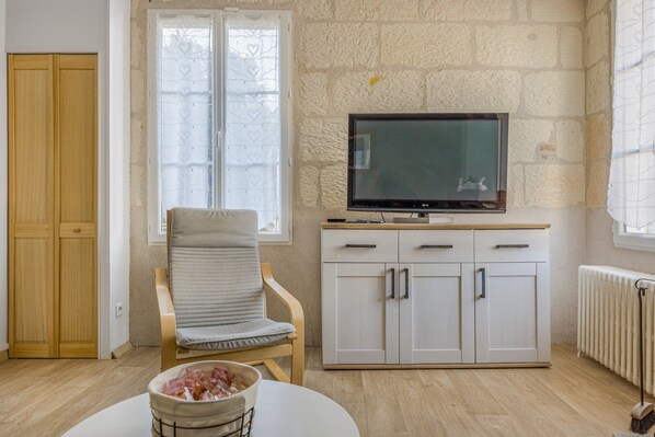 A room with a wooden wardrobe, a white cabinet with drawers, a television on top, a cushioned chair, and a radiator.