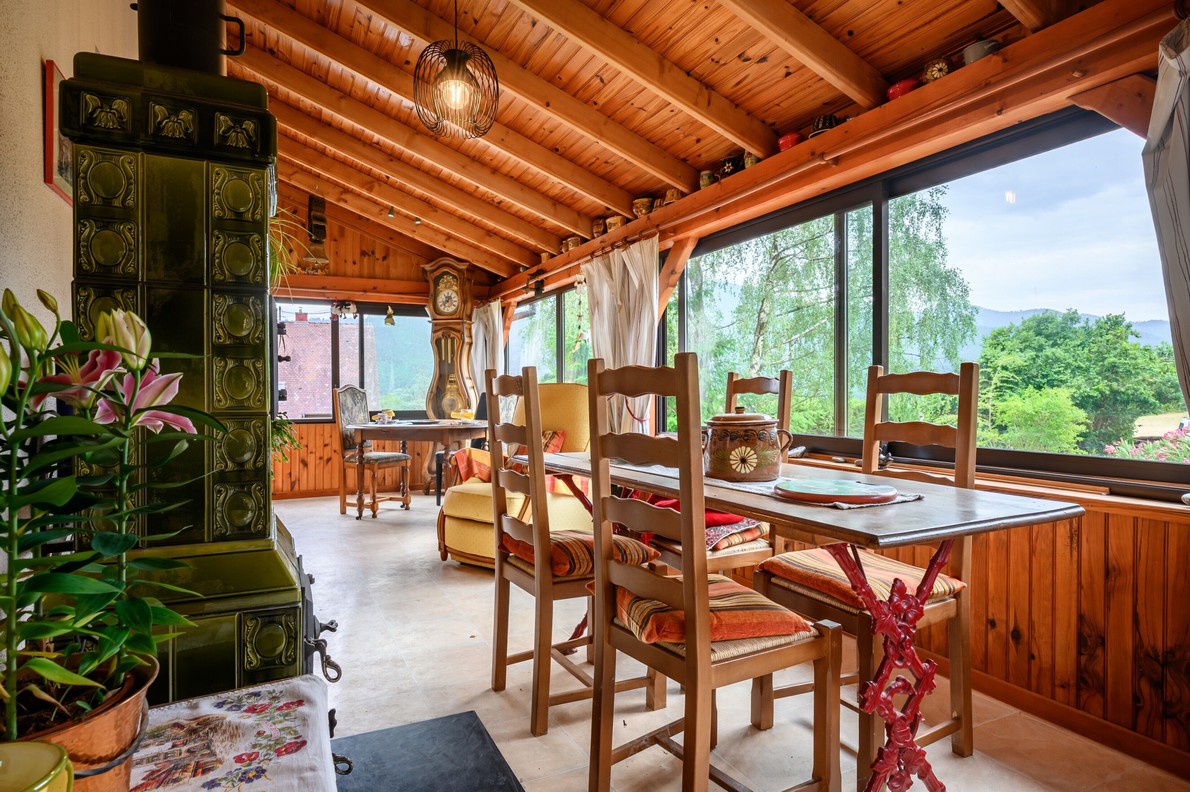 A wooden interior with a dining table, chairs, a clock, and a view of greenery outside.