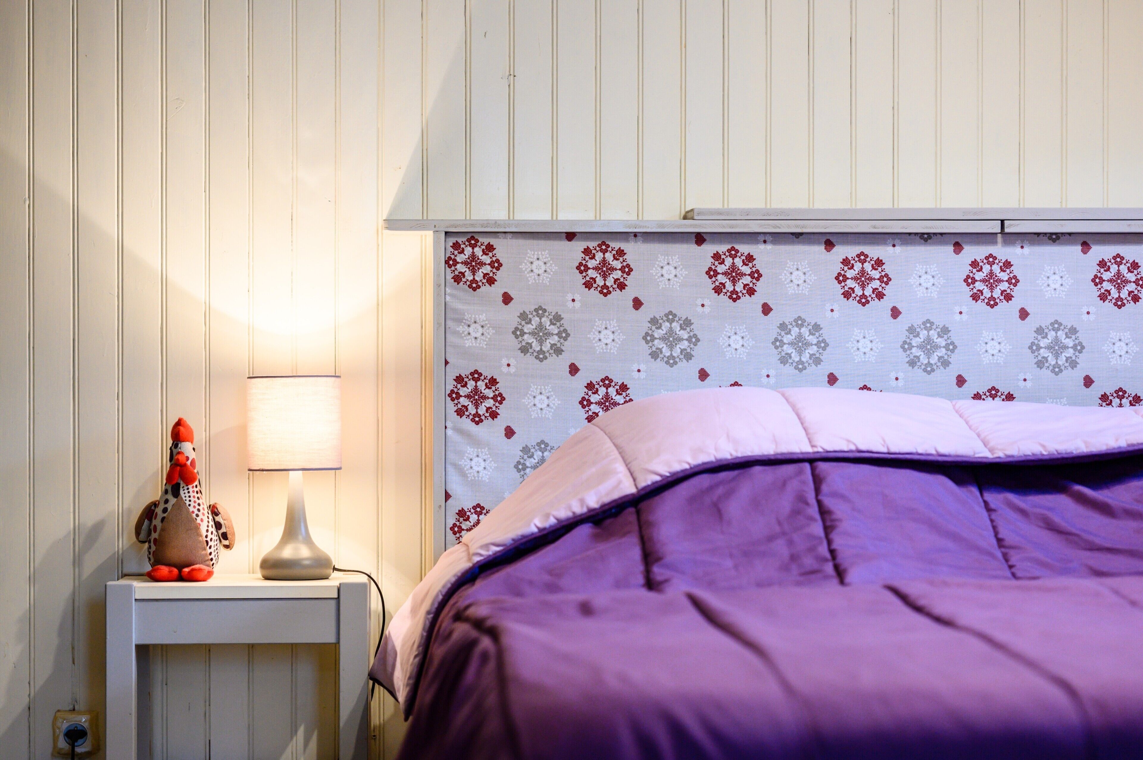 Ein Schlafzimmer mit violettem Bettbezug, einer Lampe auf einem Nachttisch und einer Wand mit floralem Muster.