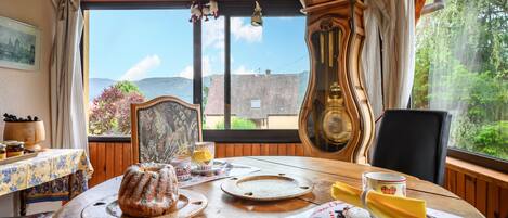 A dining table with a cake, plates, and cutlery, a clock, and a window with a view of greenery and buildings.