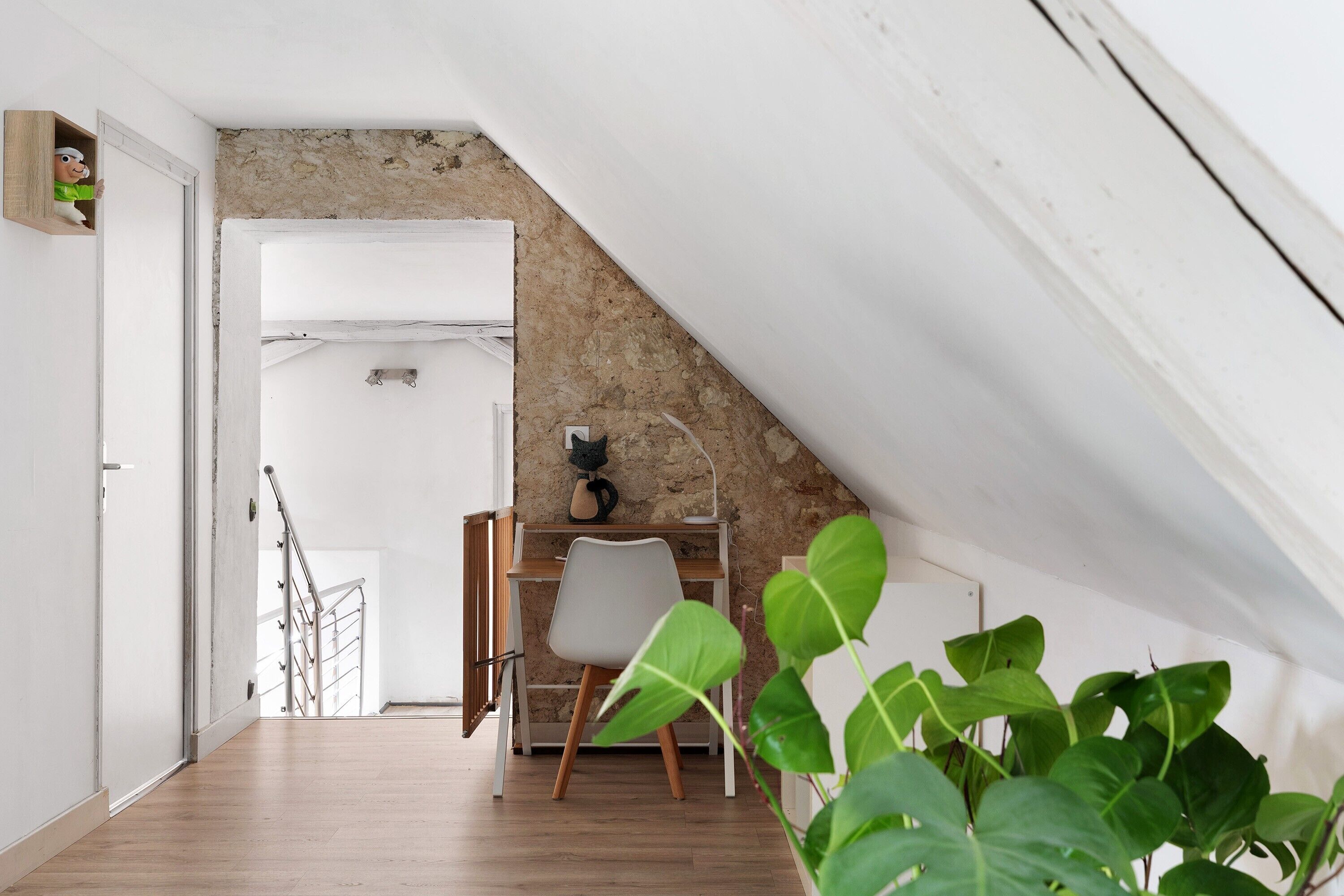 A room with a sloped ceiling, a white door, a desk with a chair, and a plant.