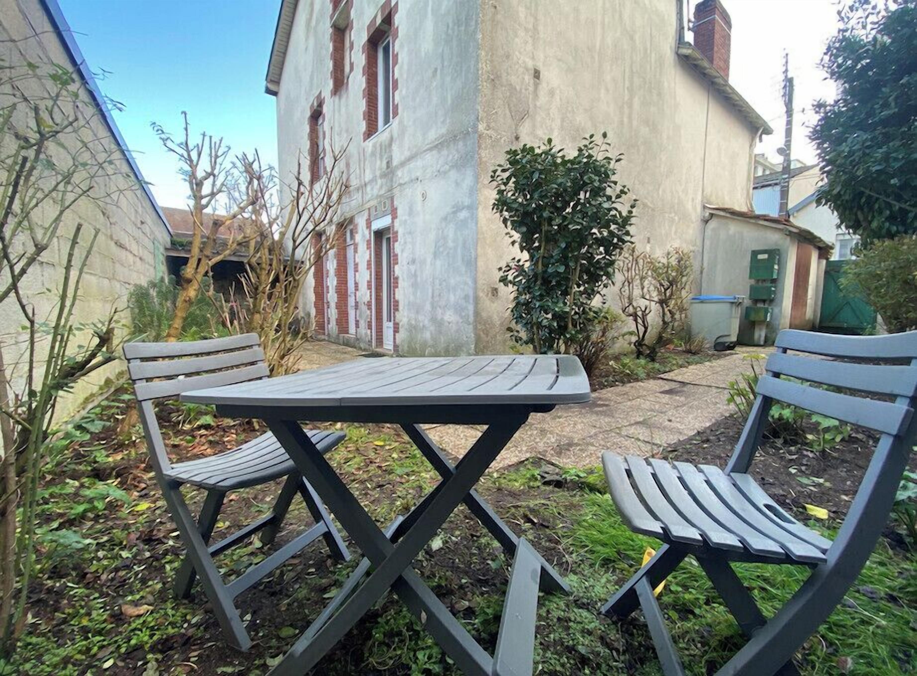 Un patio con mesa y dos sillas, rodeado de jardín y un edificio al fondo.