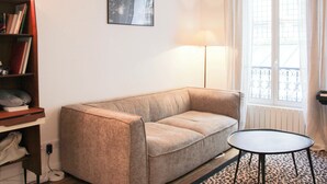 Een woonkamer met een bank, een klein rond tafeltje, een boekenkast, een lijst met een afbeelding aan de muur en een raam met gordijnen.