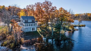 A two-story house with a large front porch, surrounded by autumn foliage and a lake. - Cozy Lakehouse with Game Room (Orono)