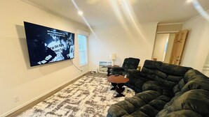 En stue med en krogsofa, et mønstret gulvtæppe, et fladskærms-tv ophængt på væggen og et lille sofabord med en lampe.