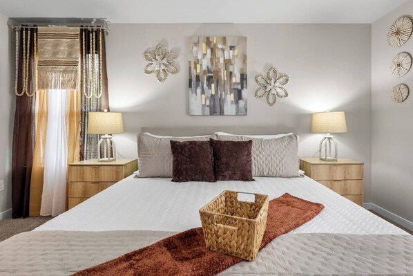 Een keurig opgemaakt hotelkamer met een groot bed, twee nachtkastjes met lampen, decoratieve wandkunst en een geweven mand op het bed.