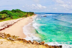 Ein Strand mit klarem türkisblauem Wasser, sandigem Ufer und einem Felsen im Vordergrund.