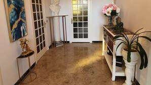 A hallway with a marble floor, a white door with a glass panel, a bust on a pedestal, a decorative ceiling light, and a console table with a vase of flowers.