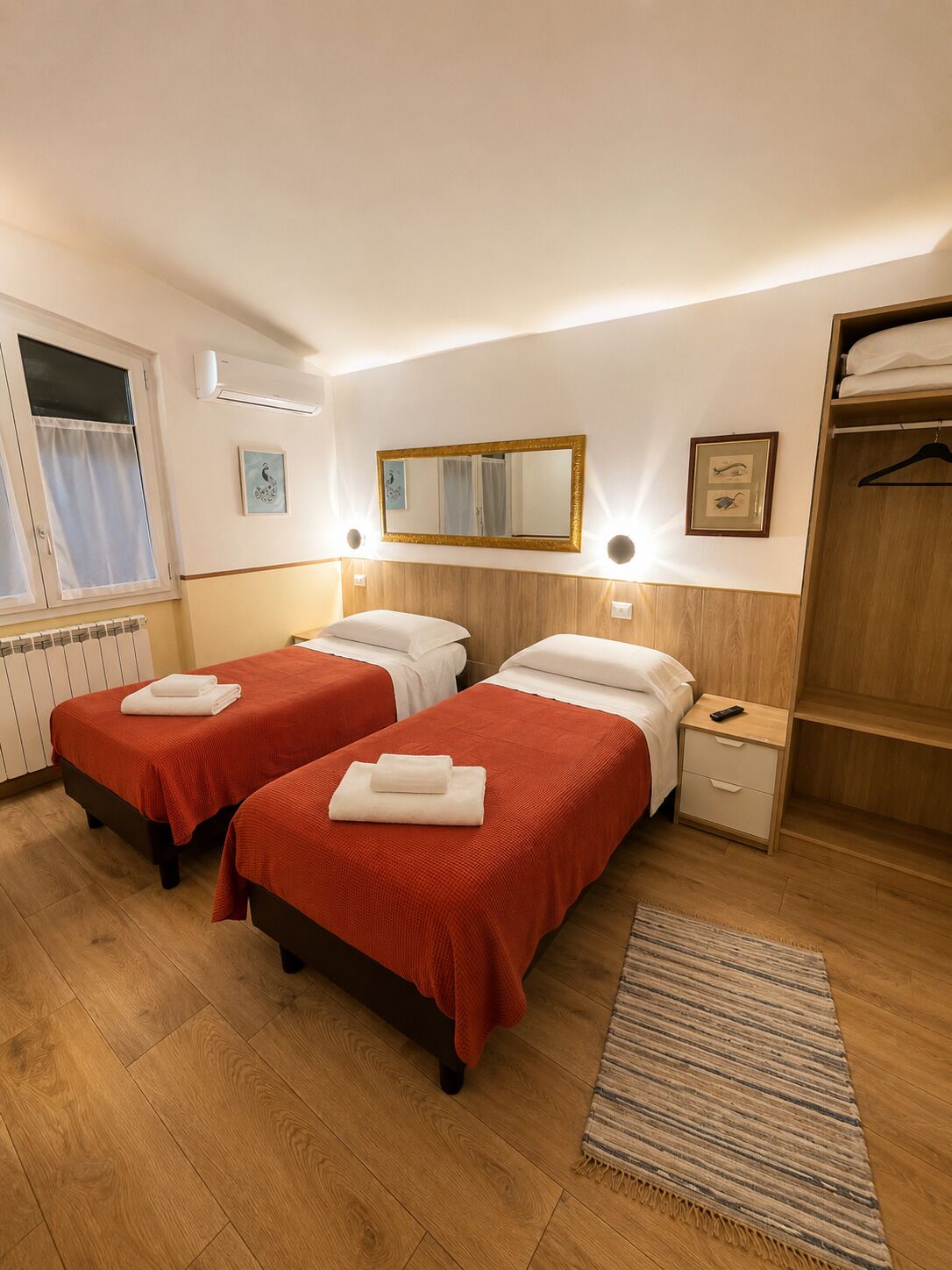 Et hotelværelse med to senge, en træskab, et natbord med lampe, et vindue med gardiner og et spejl på væggen.