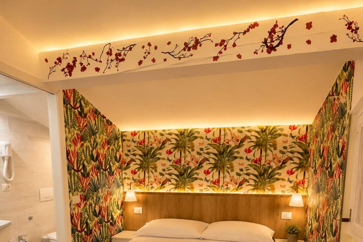 A bedroom with a bed, two nightstands, a wooden headboard, a bathroom with a bidet, and floral wallpaper.
