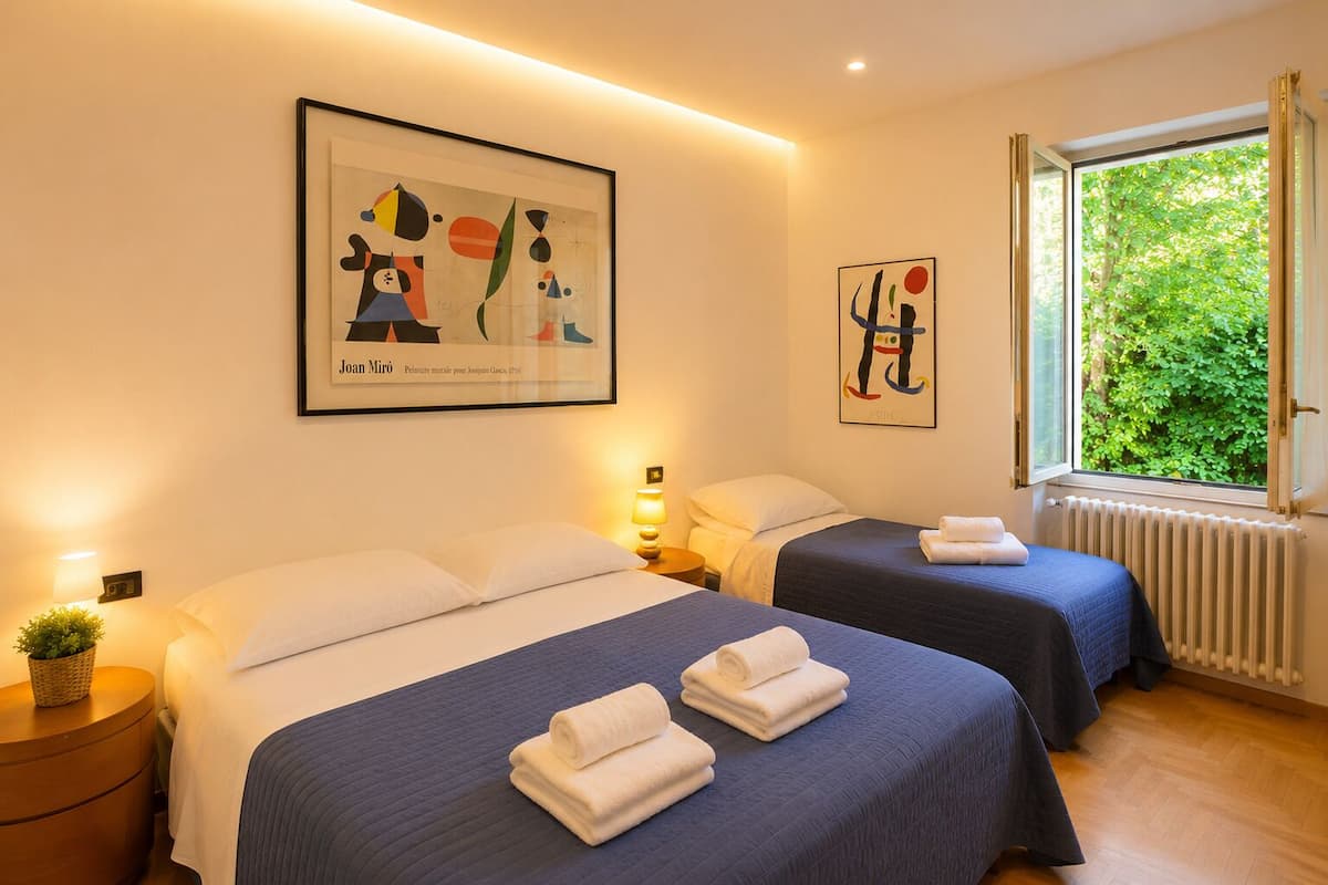 A hotel room with two beds, a large window with a view of greenery, and artwork on the walls.
