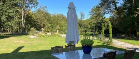 Un patio al aire libre con una mesa y sillas de vidrio, rodeado por un exuberante jardín y árboles.