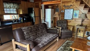 Ein gemütliches Blockhauss-Interieur mit einer Couch, Sesseln, einem Esstisch, einer Küchenzeile und einer Treppe.