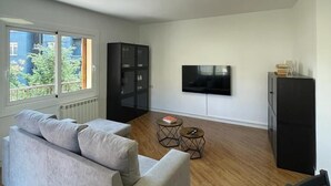 A modern living room with a grey sofa, a black cabinet, a TV mounted on the wall, and a window with a view of greenery outside. - Apartamento céntrico en Berga - ALBERGA (Berga)