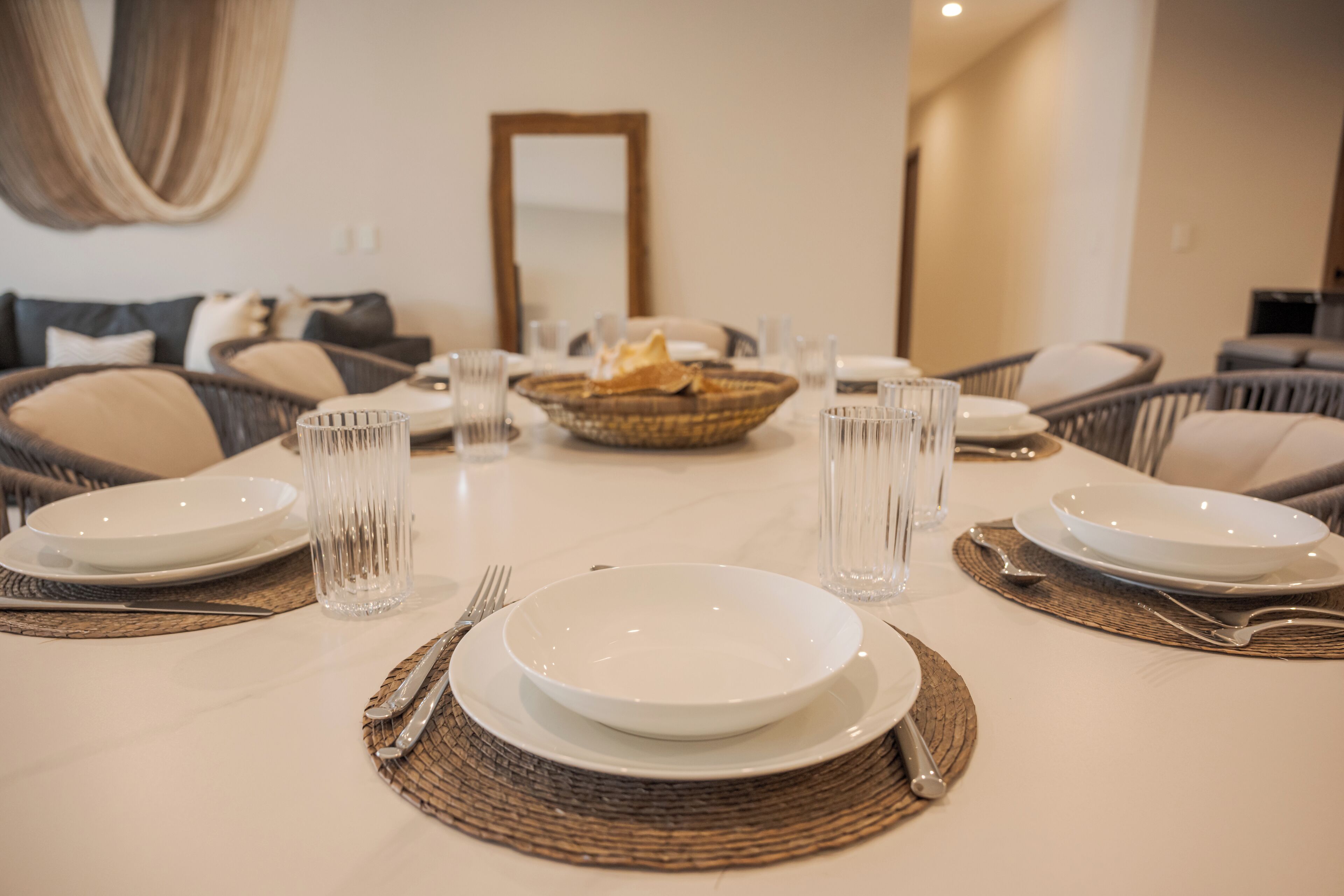 Una mesa de comedor dispuesta para seis personas, con platos blancos, vasos transparentes y un mantel tejido.