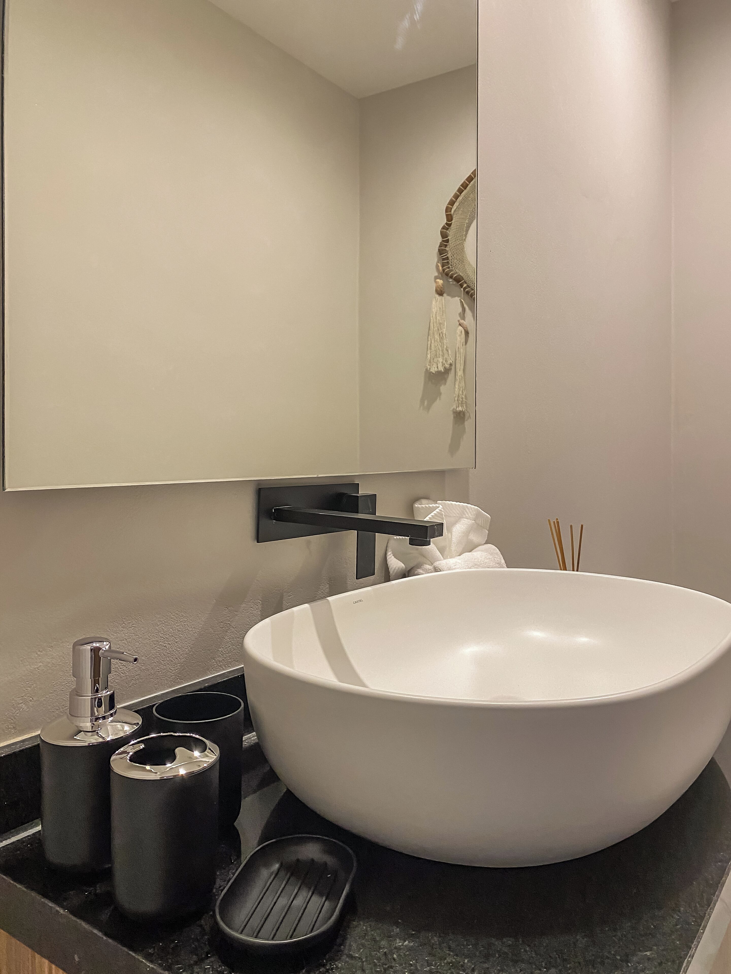 Een moderne badkamer met een grote witte wasbak, een zwarte kraan en een spiegel erboven. Op het aanrecht staan twee zwarte prullenbakken en een zwart zeepbakje.