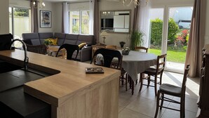 A modern kitchen with a central island, dining area with wooden chairs, a sofa, and a large window with curtains.